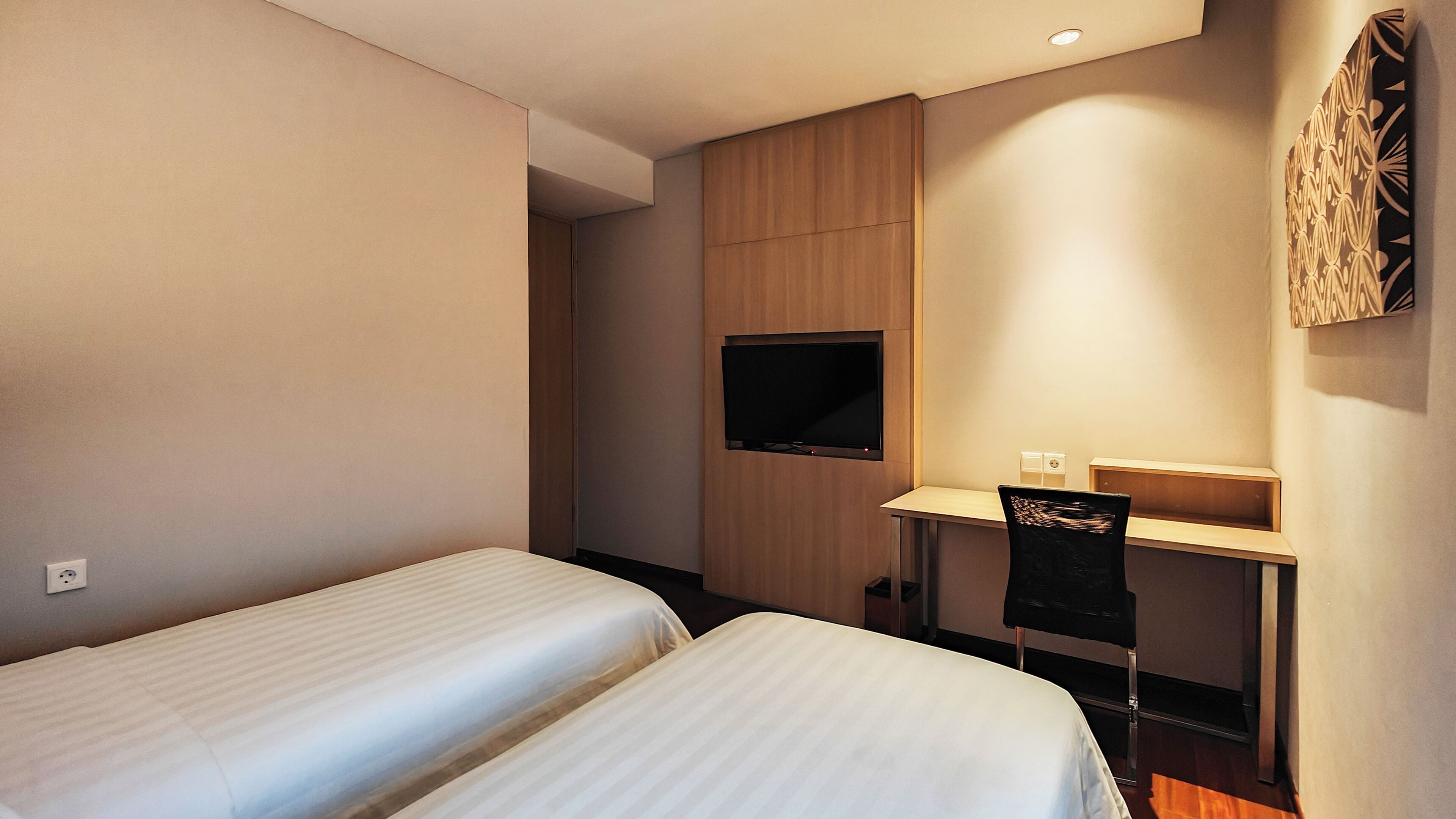 A hotel room with two single beds, a desk with a chair, a flat-screen TV mounted on the wall, and a small cabinet.