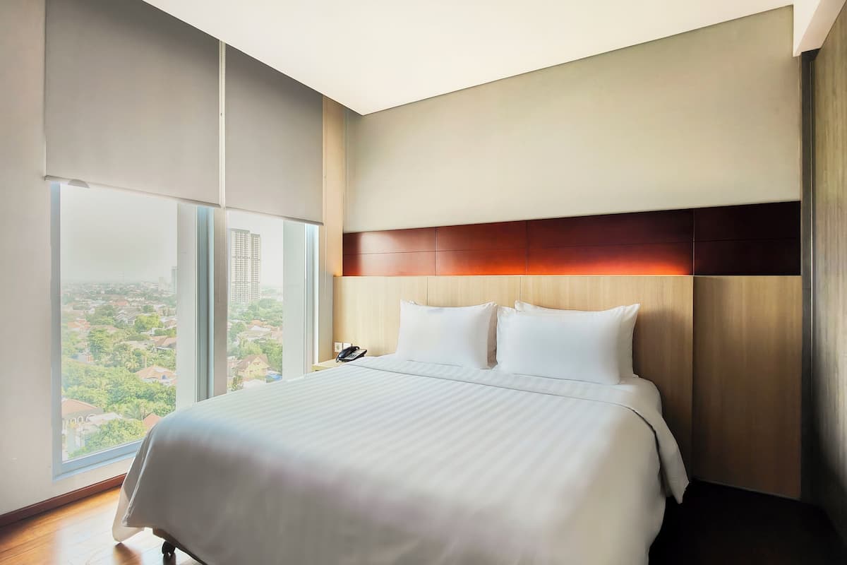 A hotel room with a large bed, a headboard with a wooden panel, a telephone on the nightstand, and a view of a cityscape through a large window.