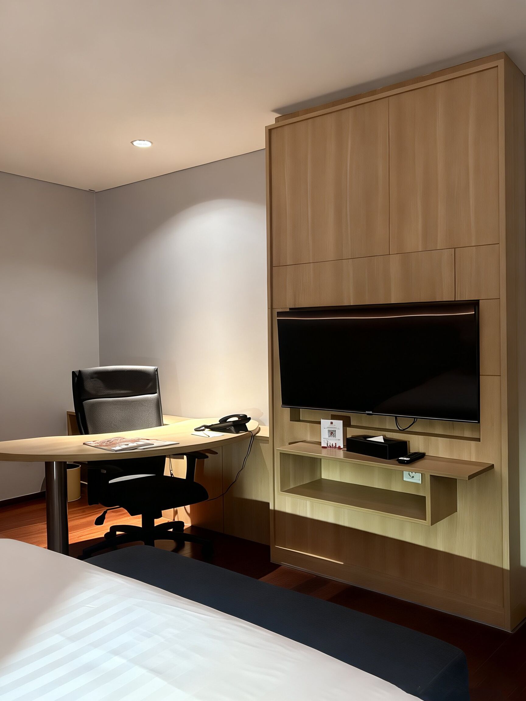 A hotel room with a bed, a desk with a chair, a flat-screen TV mounted on a wooden panel, and a small shelf below the TV.
