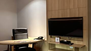 A hotel room with a bed, a desk with a chair, a flat-screen TV mounted on a wooden panel, and a small shelf below the TV.