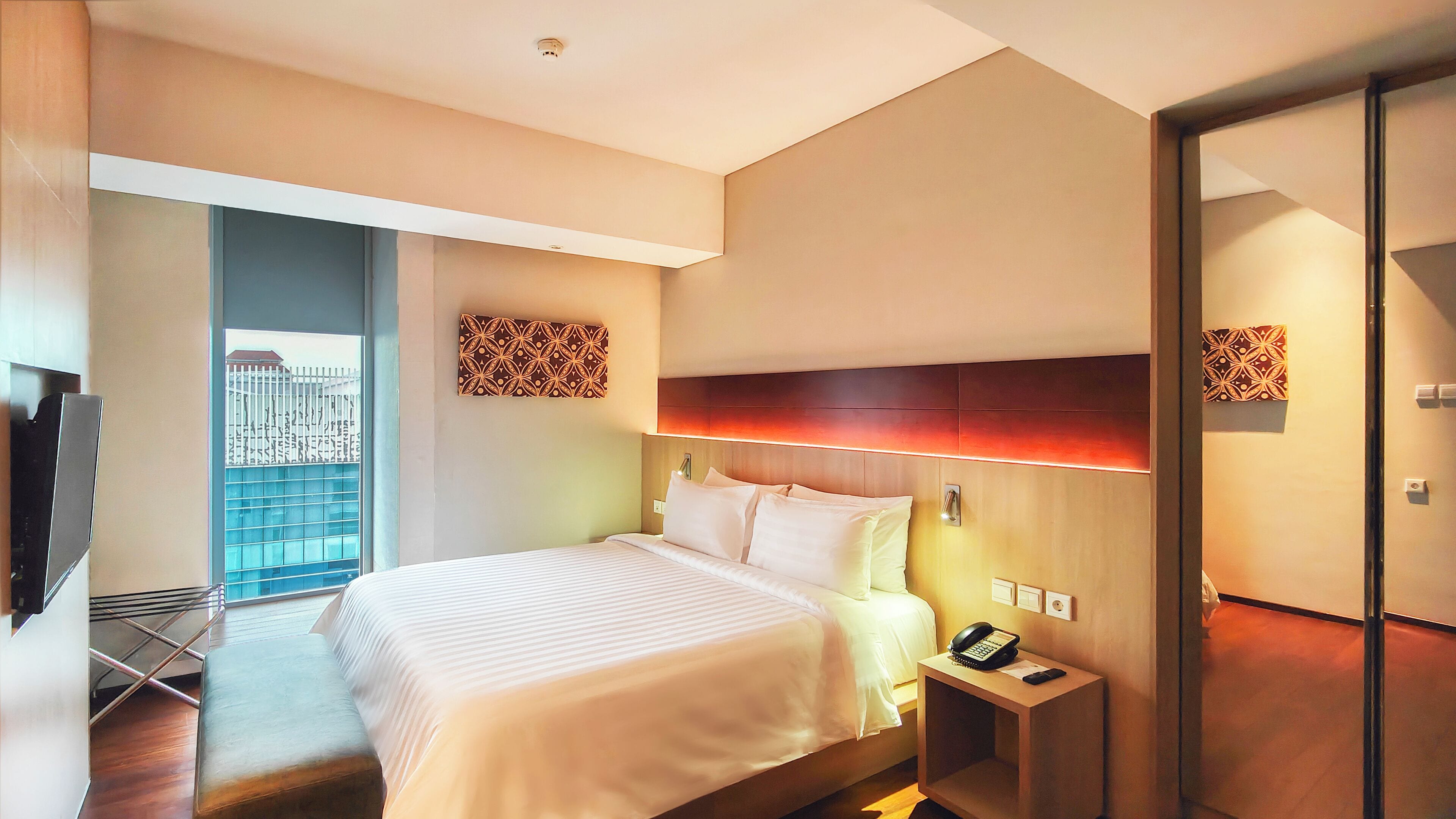 A hotel room with a large bed, a bedside table with a phone, and a view of a cityscape through a window.