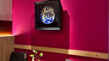 A modern room with a vibrant red wall, a framed artwork, two black chairs, a small round table with a vase and a bottle, and a wooden panel running horizontally.