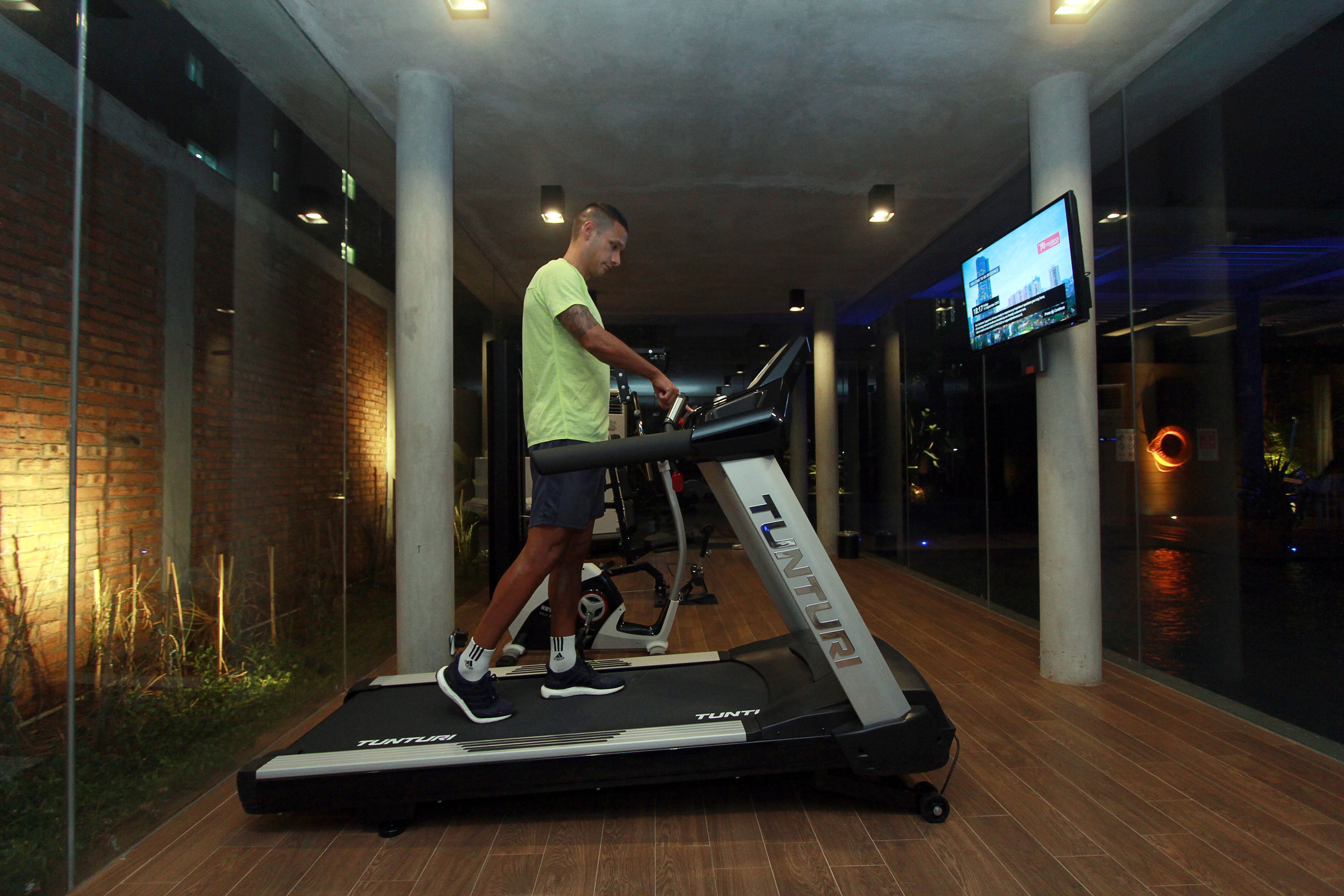A man using a treadmill in a modern indoor gym with brick walls and a television screen on the wall.