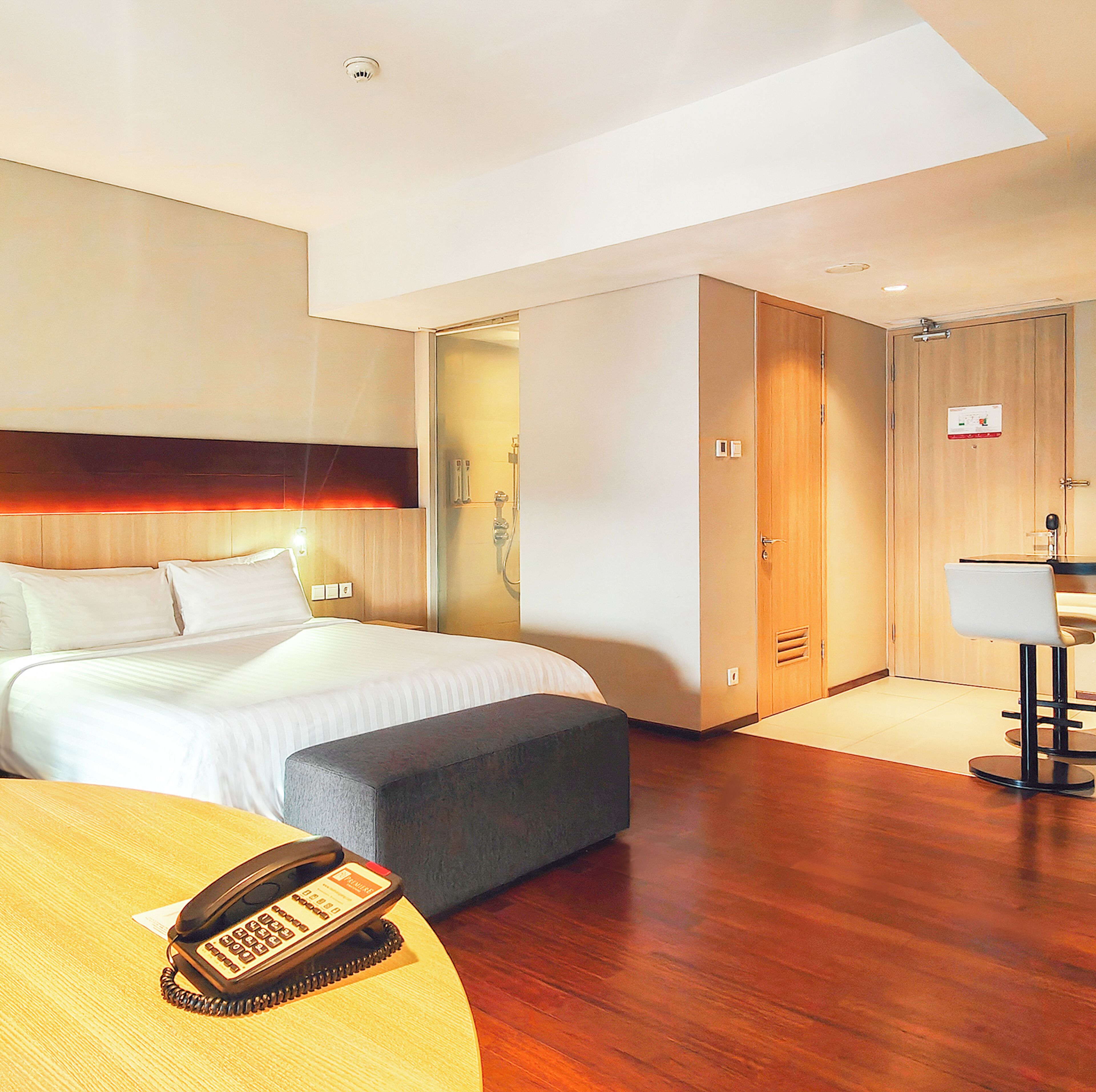 A hotel room with a bed, bedside table with a telephone, a small bench, a bathroom with a shower, and a desk with a chair.