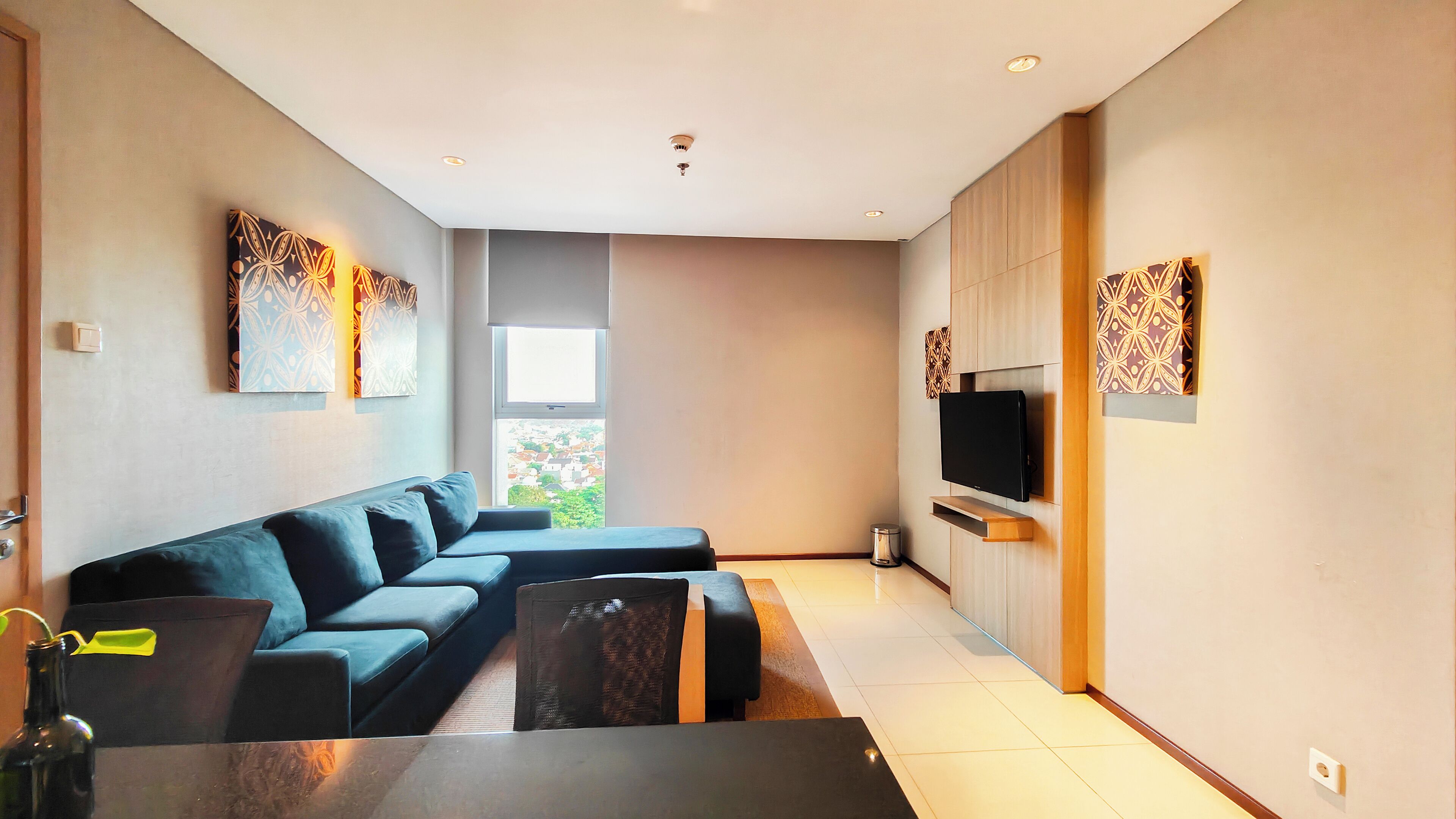 A modern living room with a sectional sofa, a flat-screen TV mounted on a wooden wall, and a large window providing a view of the city.