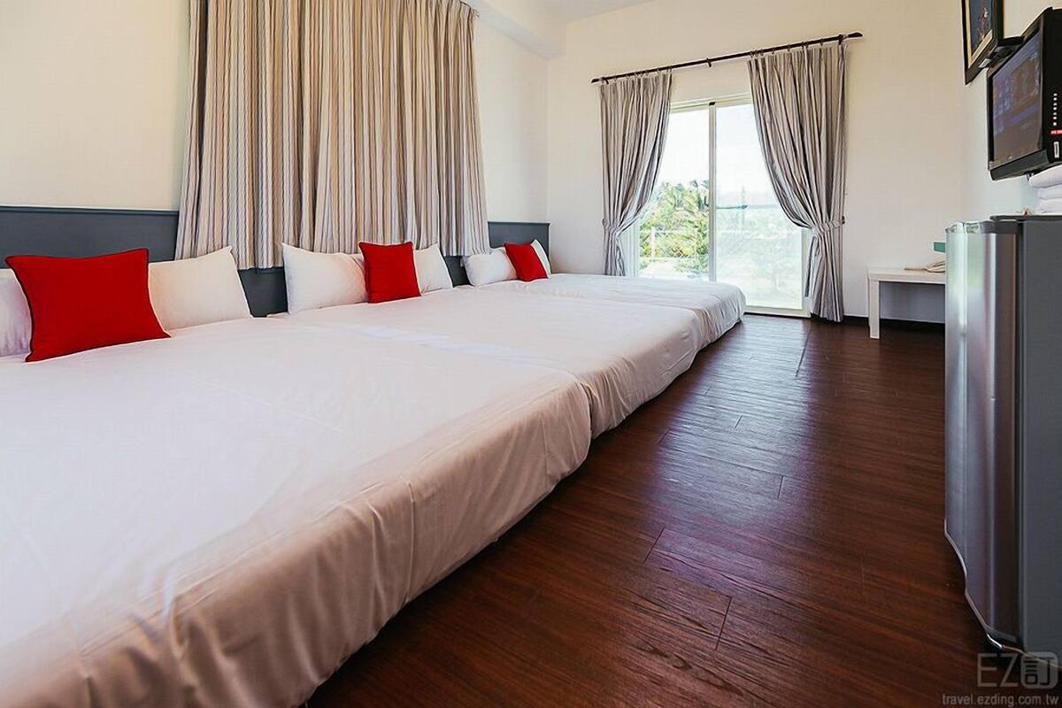 A large bed with white and red pillows, wooden flooring, and a television on a stand.