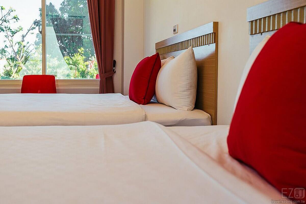 A hotel room with a large bed, two red pillows, and a view of greenery through the window.