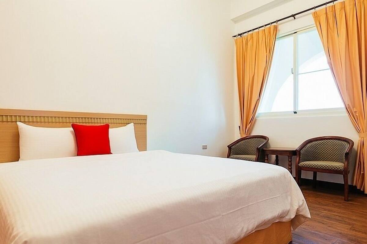 A hotel room with a large bed, two chairs, a small table, and a window with curtains.
