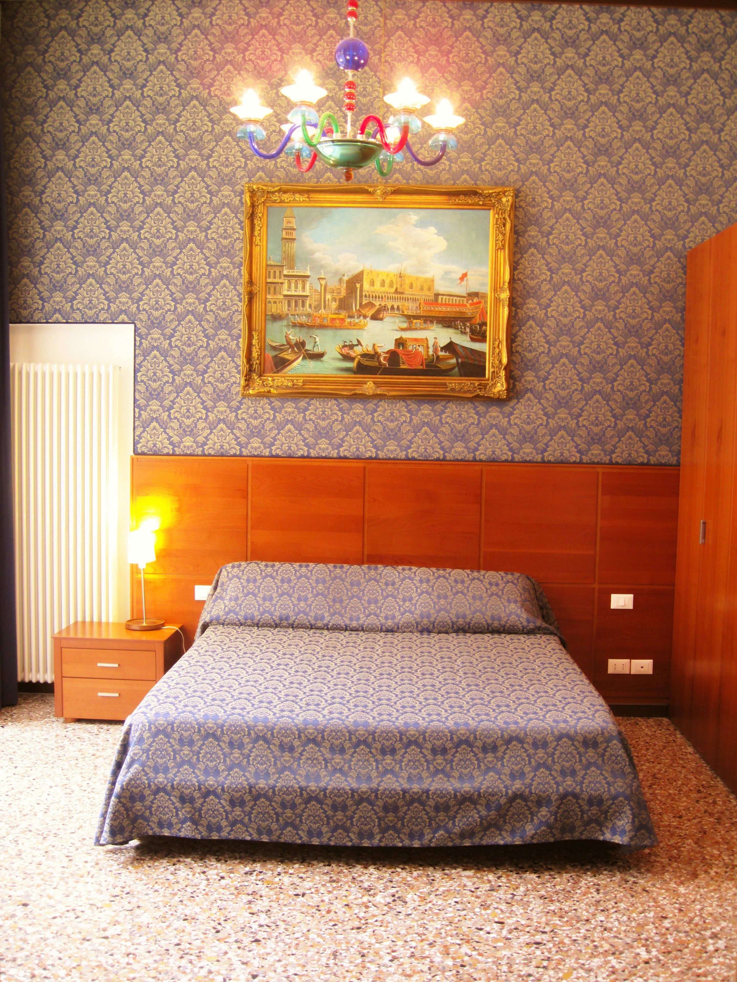 Une chambre d’hôtel avec un lit, une table de chevet, une lampe, un tableau représentant un paysage urbain et un lustre.