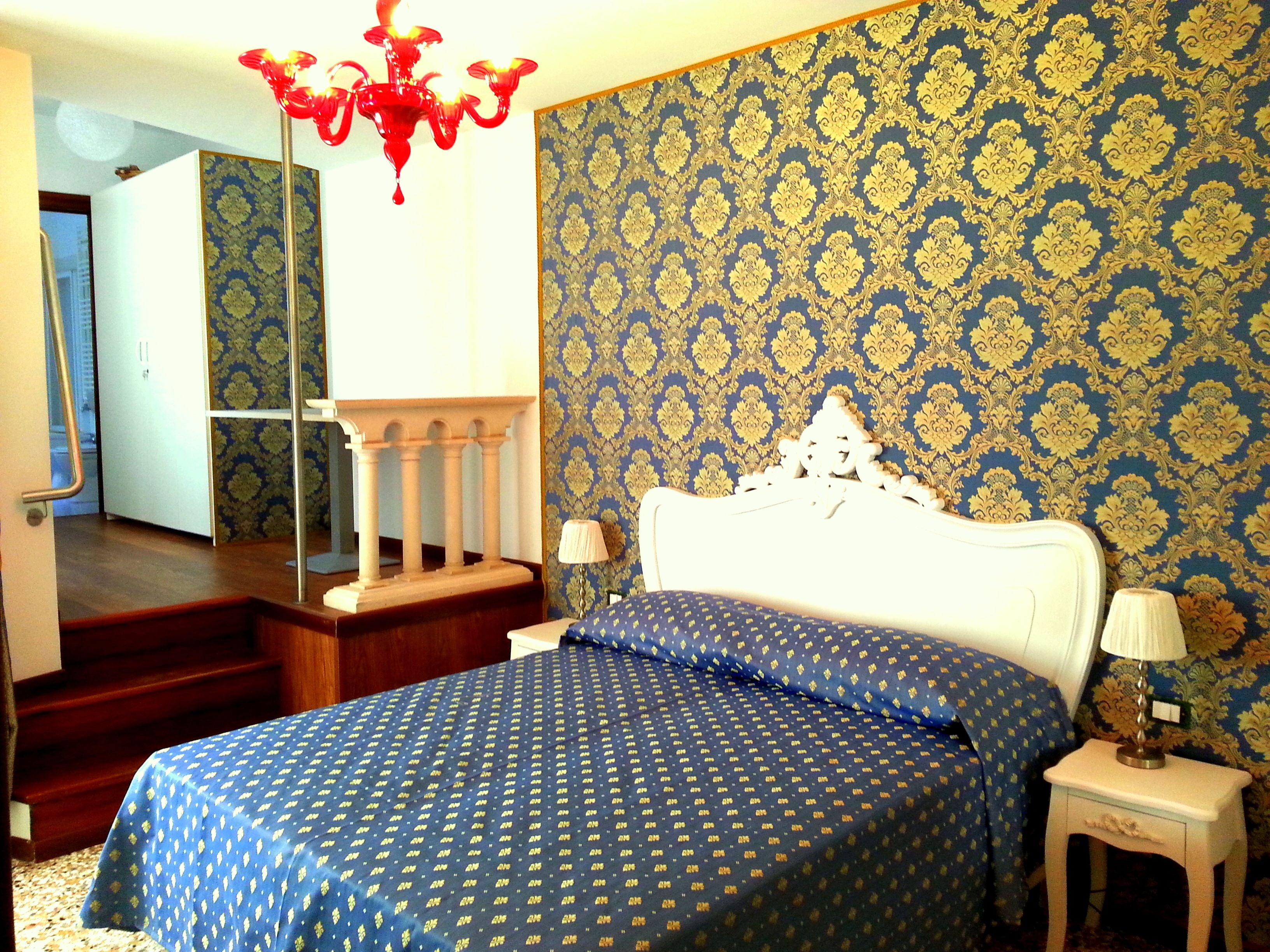 A bedroom with a patterned wallpaper, a chandelier, a bed with a patterned bedspread, a nightstand, and a lamp.