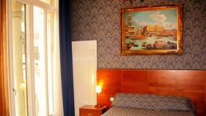 A hotel room with a bed, a nightstand, a lamp, a window with curtains, and a painting on the wall.