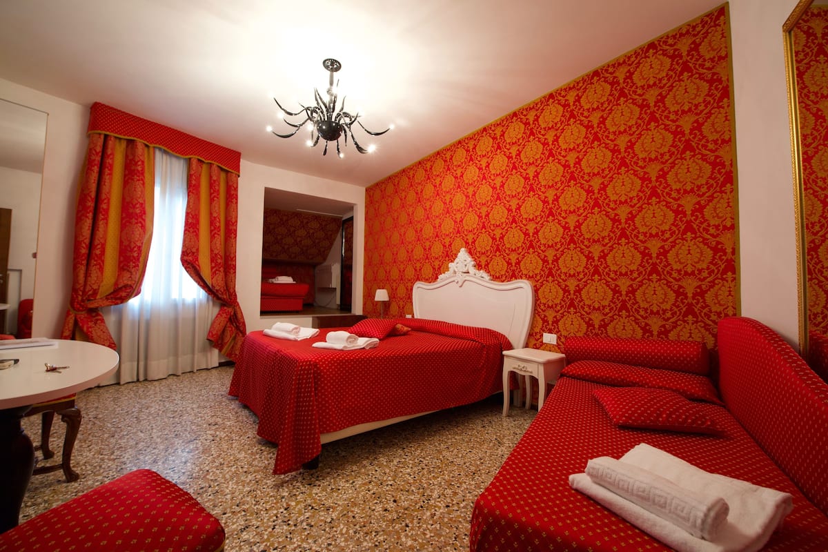 A hotel room with a red and gold patterned wall, a white bed, a red sofa, and a small round table.