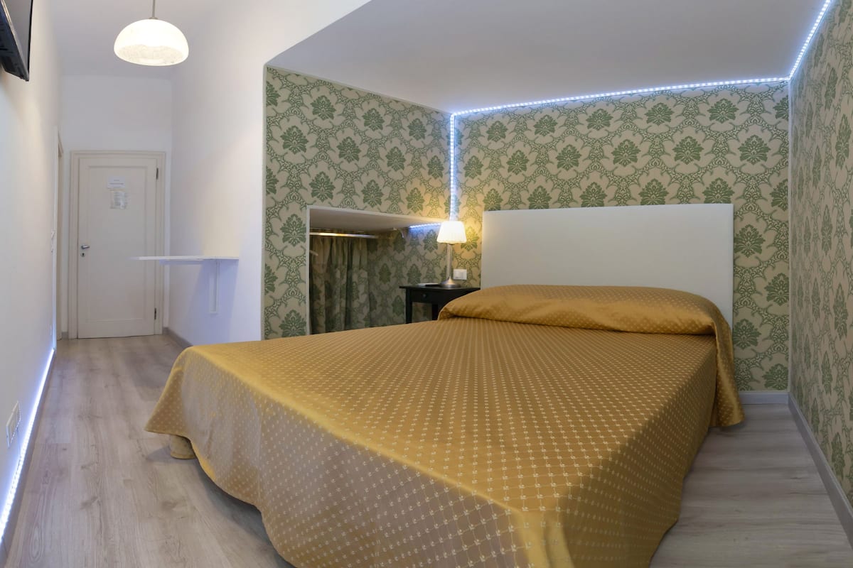 A bedroom with a bed, a nightstand, a lamp, and patterned wallpaper.