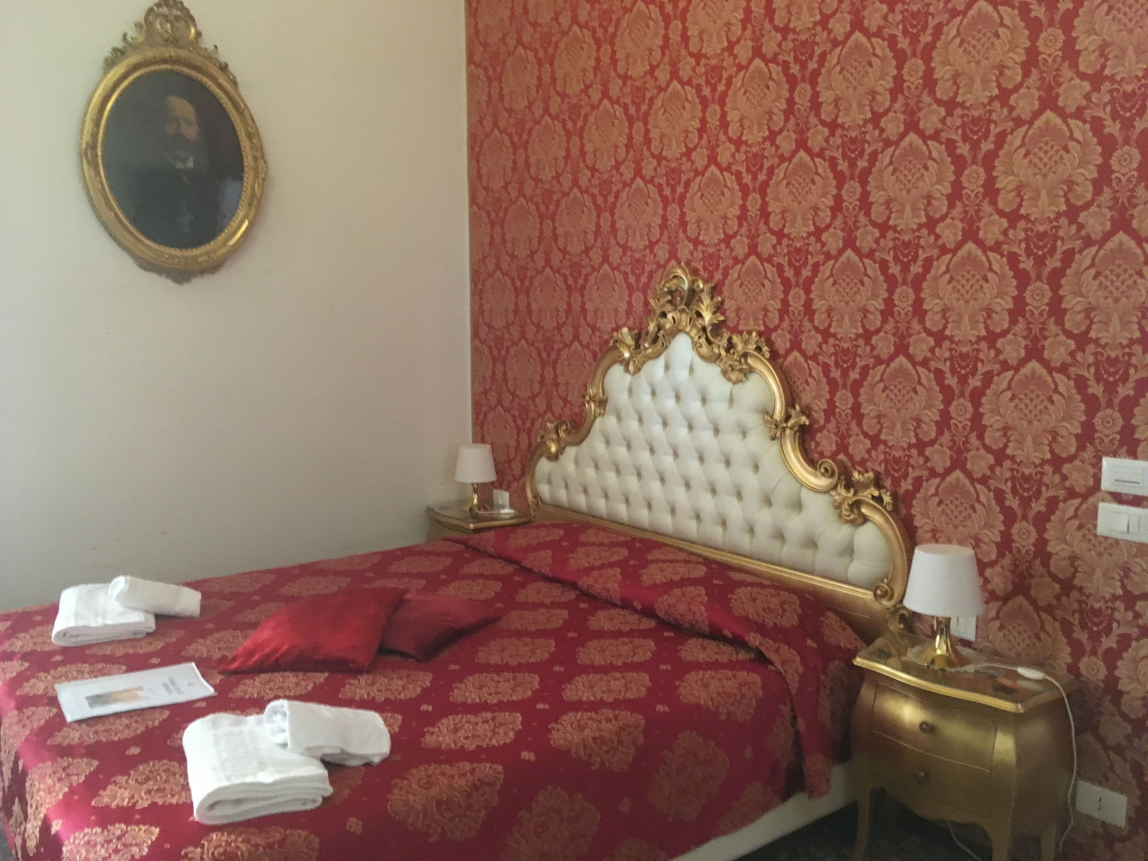 A hotel room with a red patterned wall, a white tufted headboard, and a gold-framed mirror. There are two bedside tables with lamps, a red bedspread, and neatly folded towels.