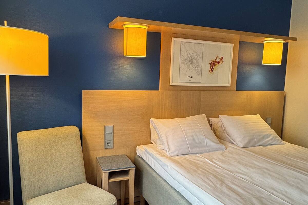 A hotel room with a bed, a chair, a small table, and two wall-mounted lamps.