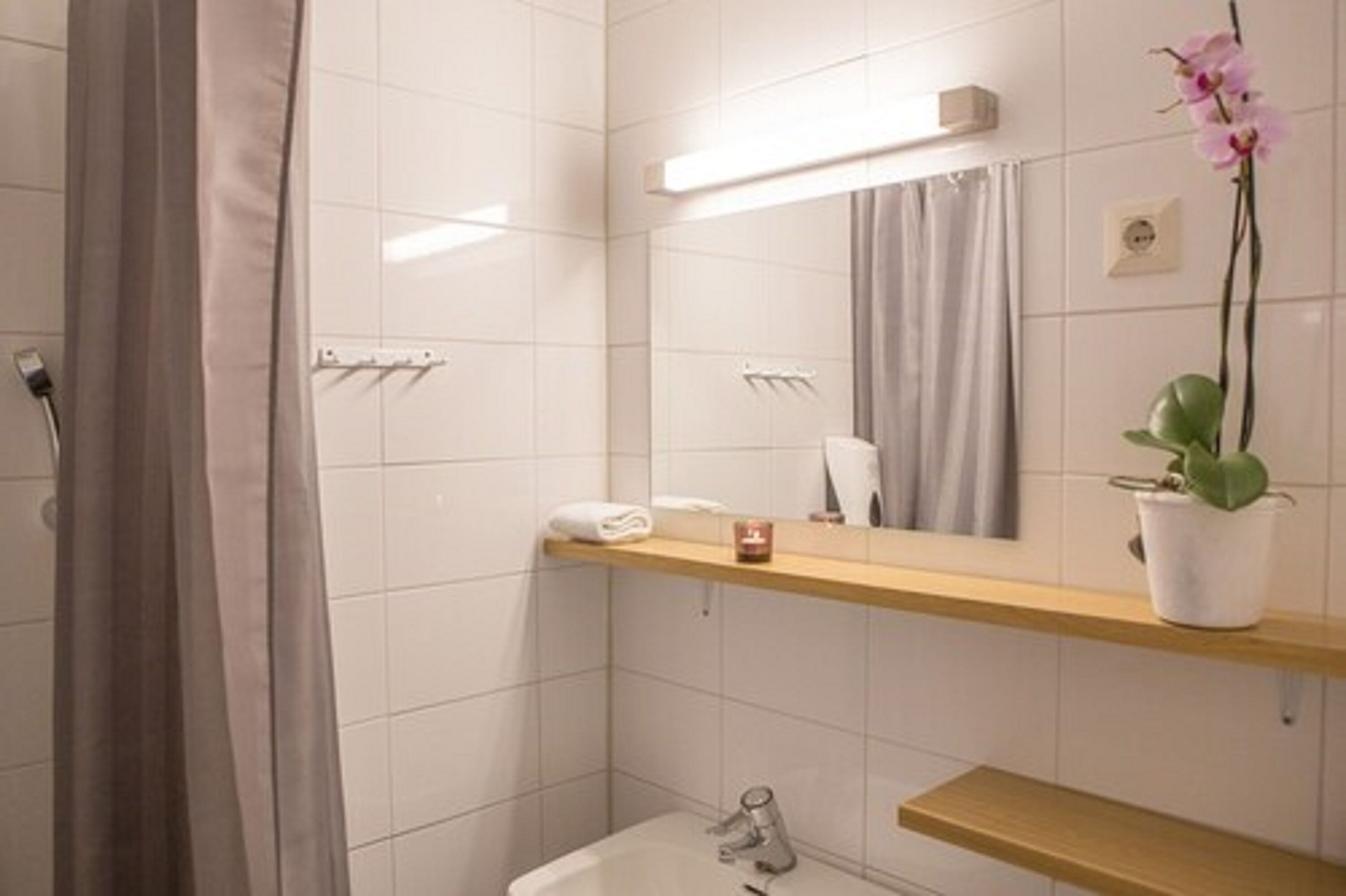 Une salle de bain moderne avec des carreaux blancs, un meuble-lavabo en bois, un miroir, un porte-serviettes et une orchidée en pot.