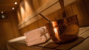 A copper bowl with a handle on a wooden bench, a folded white towel, and a wooden wall in the background. - Hotel Amado (Pori)