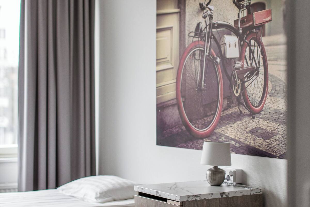 A hotel room with two beds, a nightstand with a lamp, and a large framed picture of a bicycle on the wall.