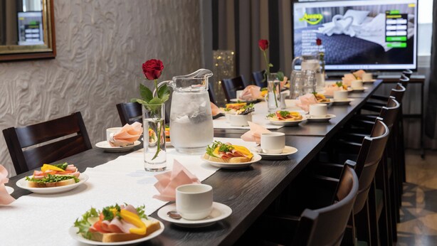 A long dining table set with plates of food, glasses, and pitchers of water, with a television screen in the background displaying a bed image. - Hotel Amado (Pori)