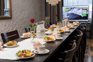 A long dining table set with plates of food, glasses, and pitchers of water, with a television screen in the background displaying a bed image. - Hotel Amado (Pori)