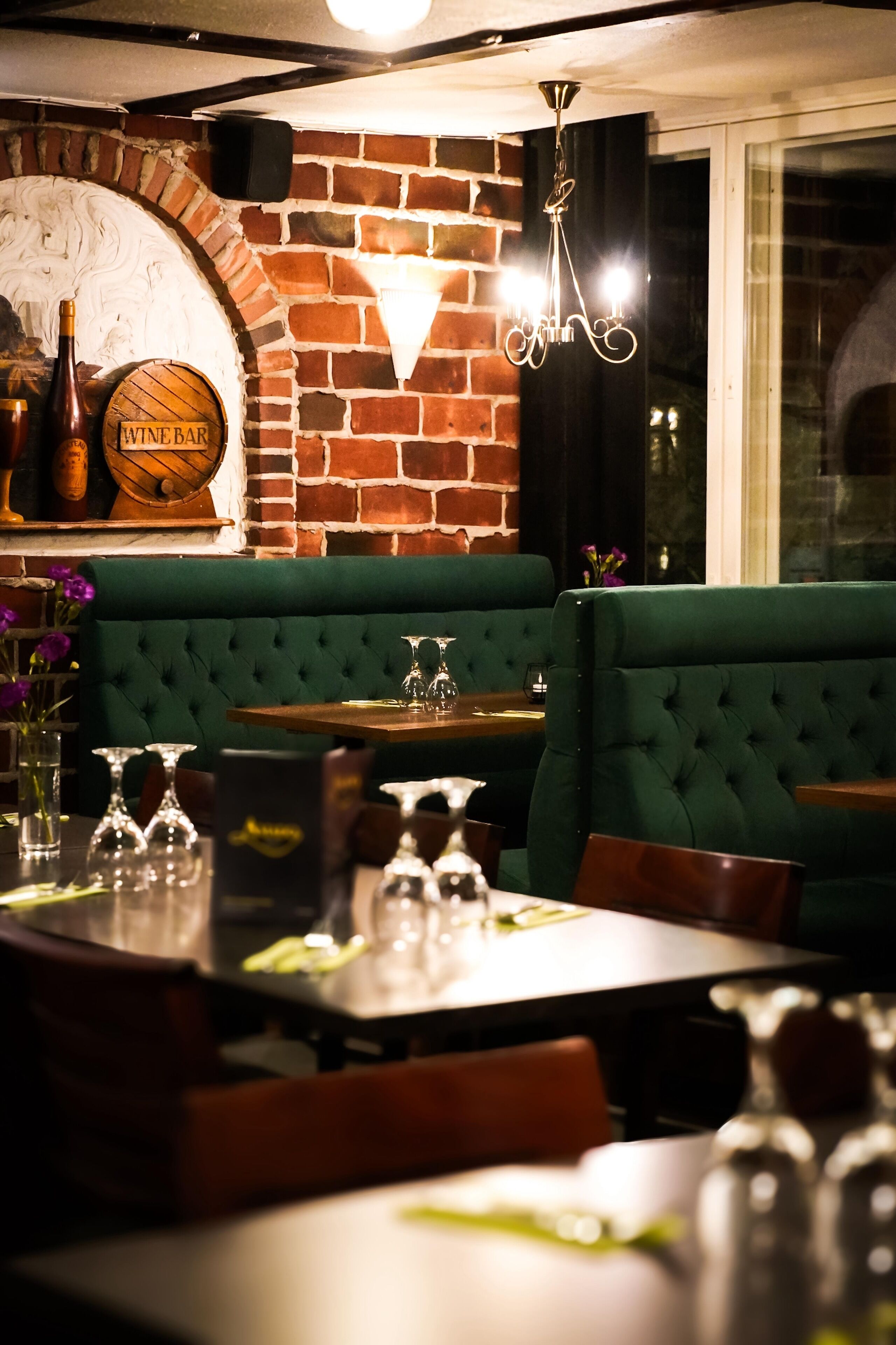 Un restaurant aux murs de briques, avec des banquettes recouvertes d’un tissu vert et des tables dressées avec des verres à vin et des couverts.