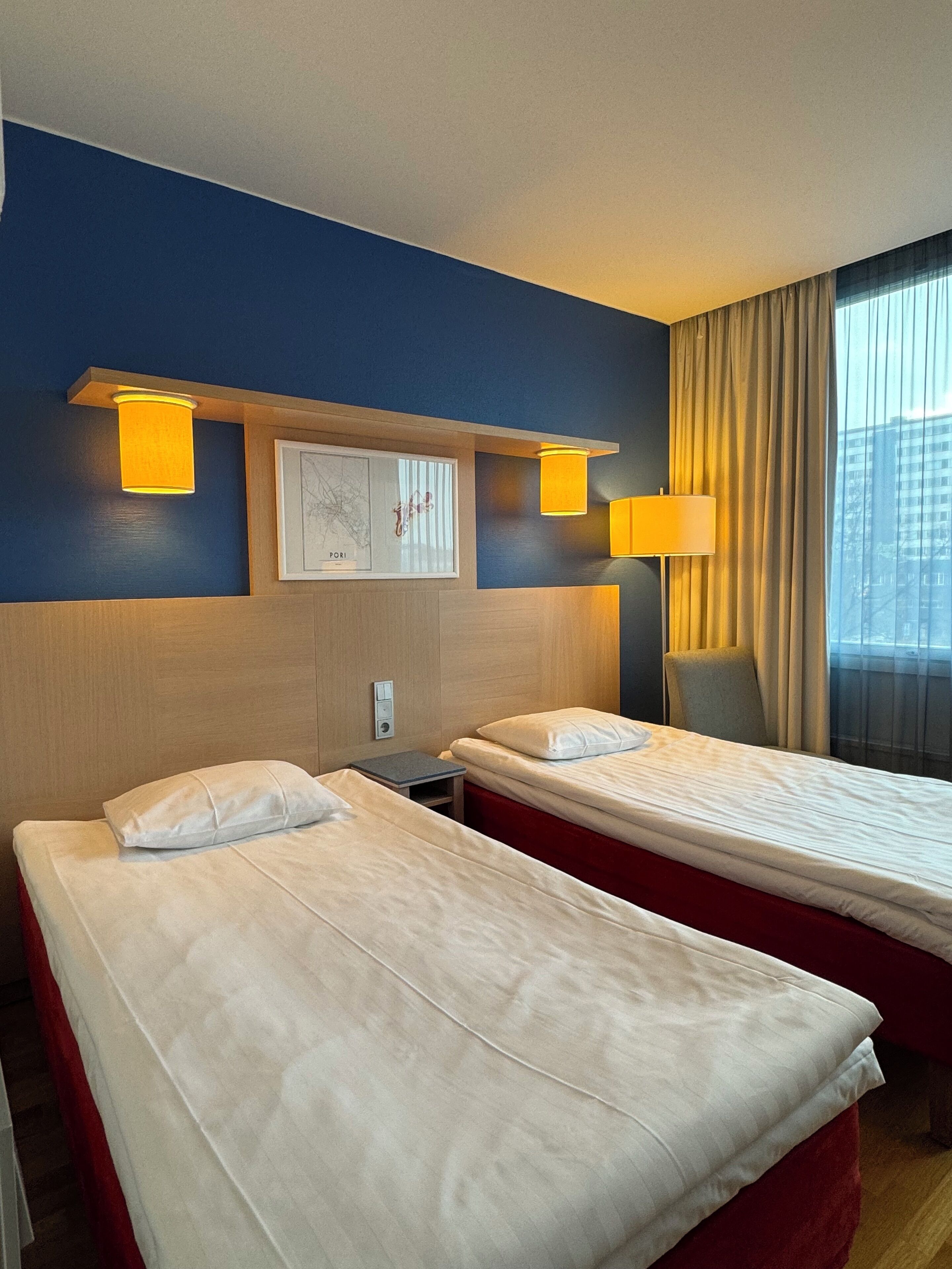 A hotel room with two single beds, a wooden headboard, a framed map, and a window with curtains.