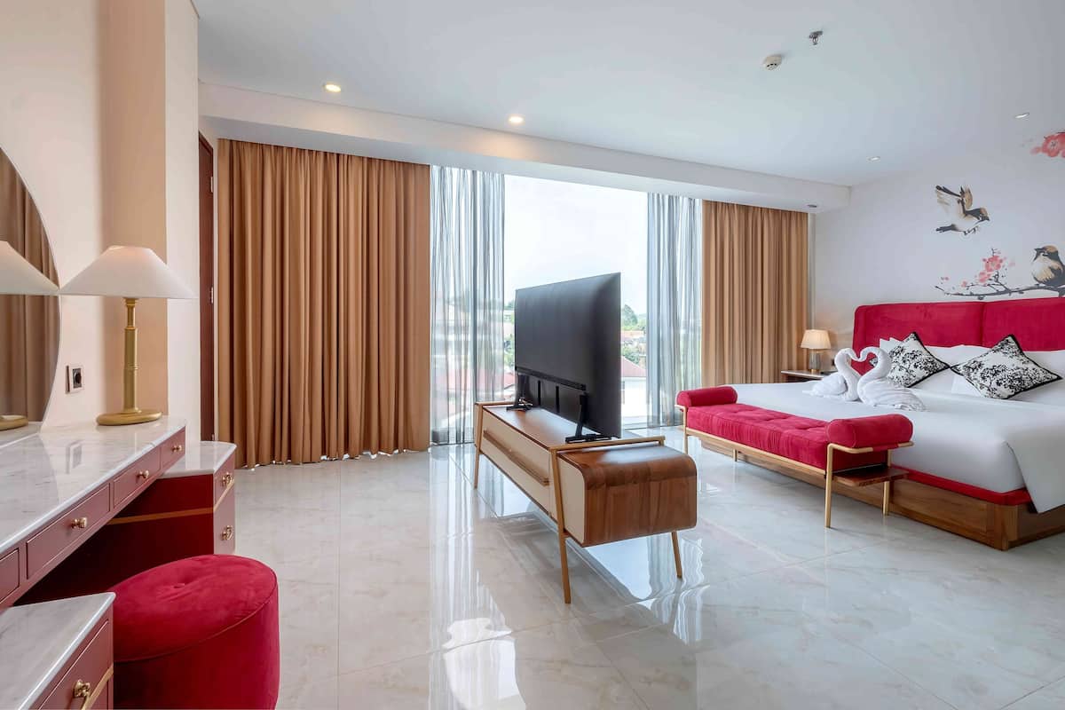 A hotel room with a bed, a TV, a desk, and a red ottoman.