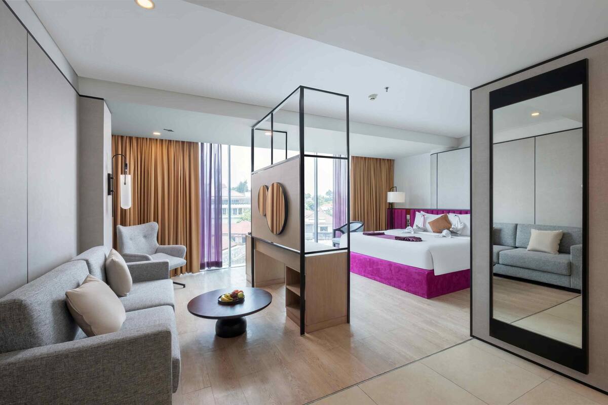 A modern hotel room with a sofa, a round table with fruit, a bed with a pink bedspread, and a large mirror.