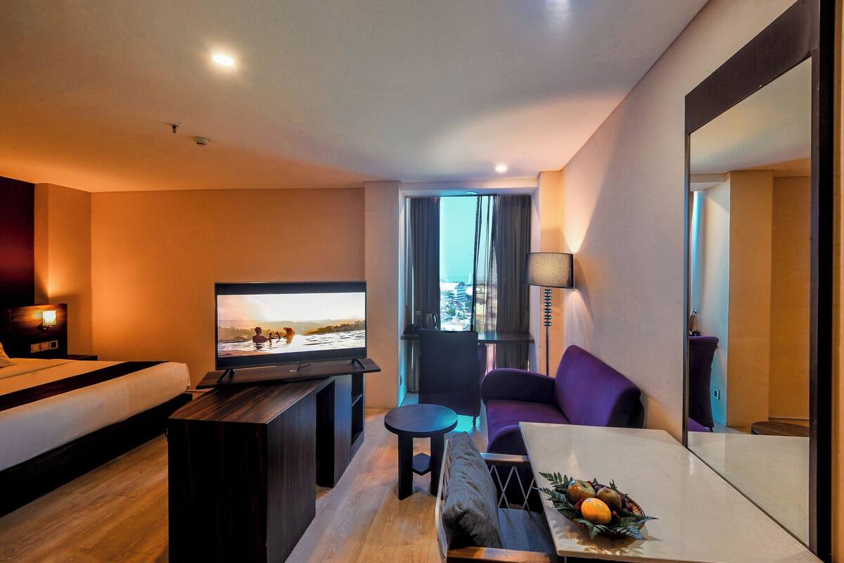 A modern hotel room with a bed, a TV, a sofa, a small table, and a view of a cityscape through the window.