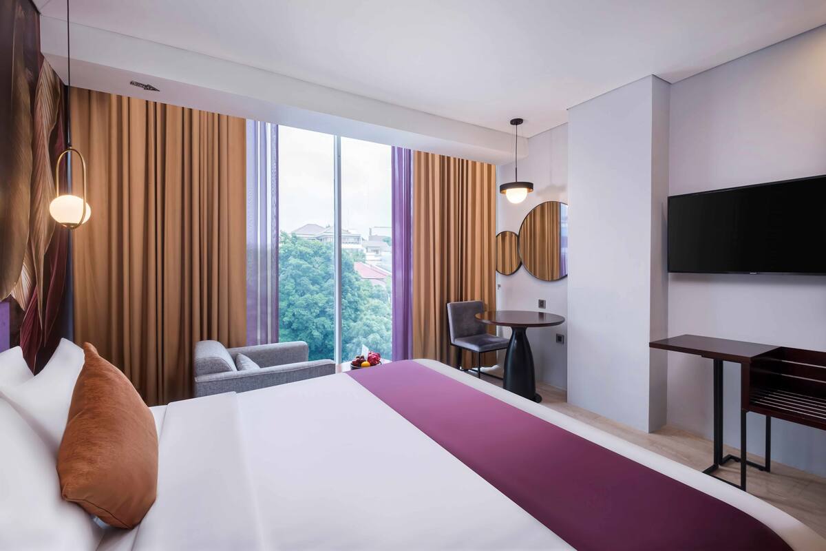 A hotel room with a large bed, a desk, a chair, a TV, and a view of the city through large windows.
