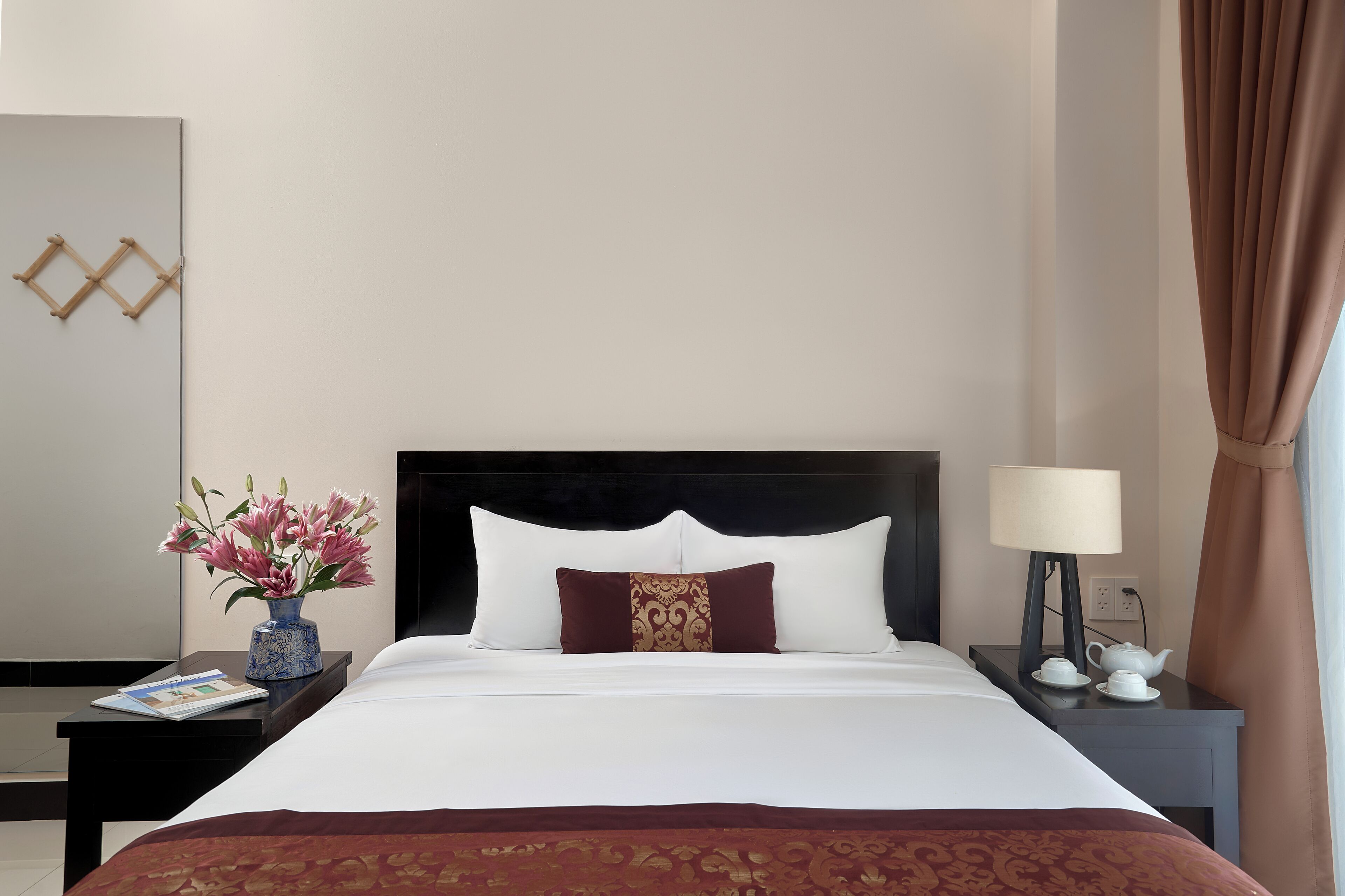 Quarto de hotel com cama, mesa de cabeceira com abajur, um vaso de flores e um objeto decorativo na parede.