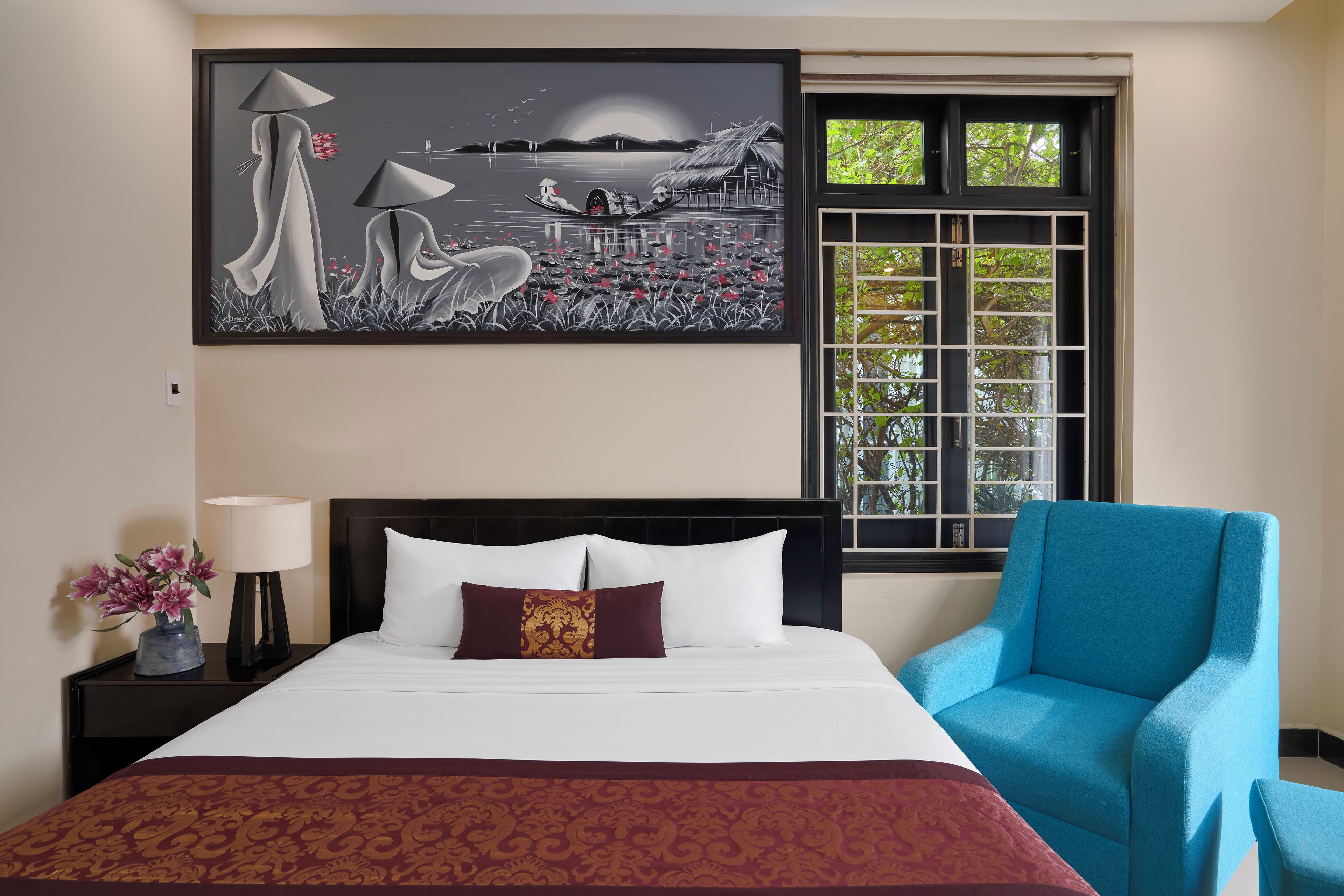 A hotel room with a bed, a blue armchair, a nightstand with a lamp, a vase of flowers, and a large painting on the wall.