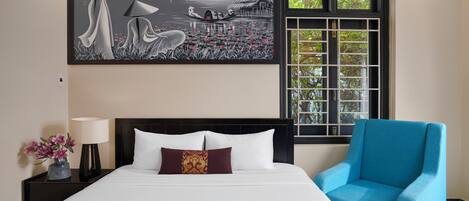 Una habitación de hotel con una cama, un sillón azul, una mesita de noche con una lámpara, un jarrón de flores y un cuadro grande en la pared.