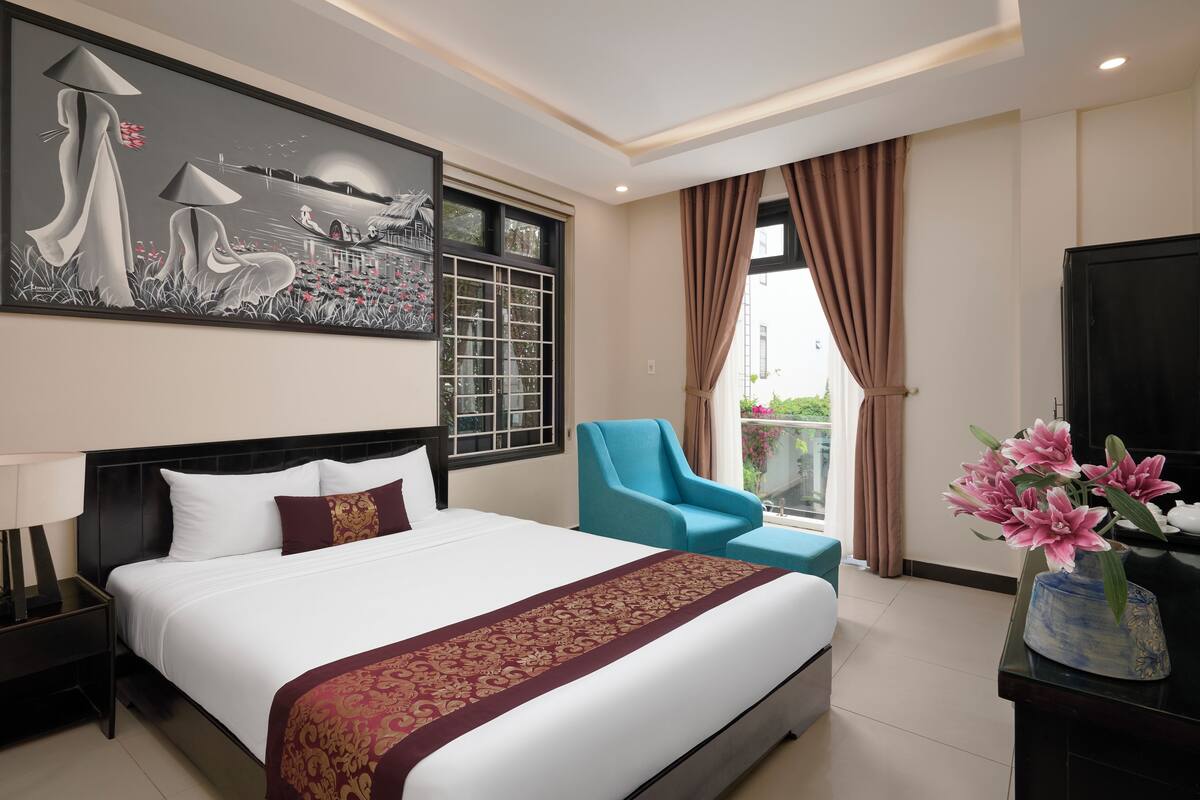 A hotel room with a large bed, a blue armchair, a painting on the wall, and a view of the outdoors.