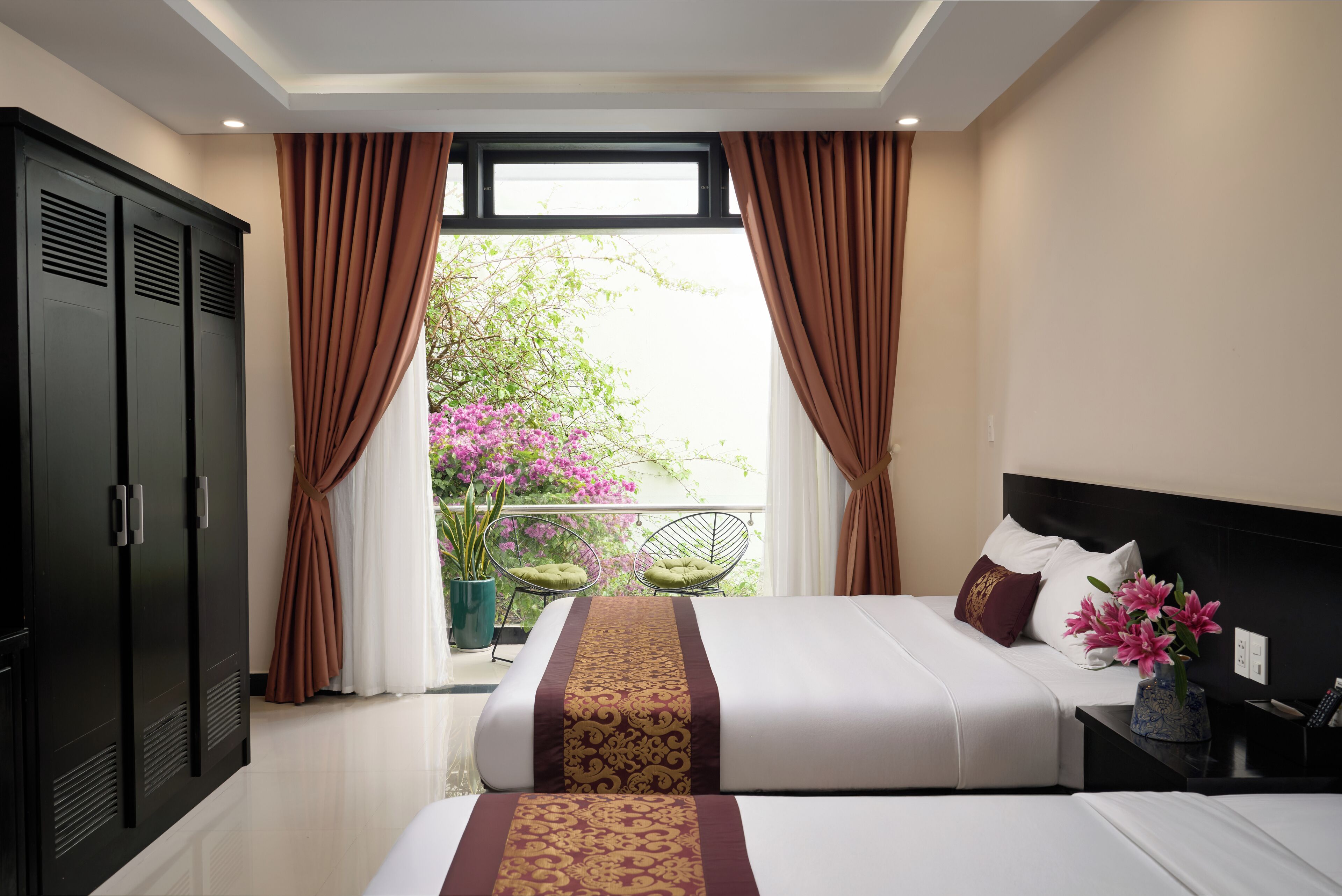 A hotel room with a large bed, a nightstand, a potted plant, and a view of a balcony with flowers.