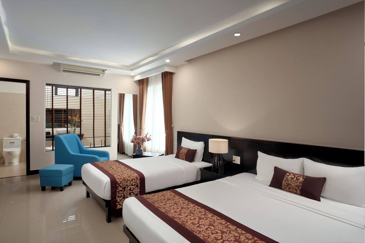 A hotel room with two beds, a blue chair, a bedside table with a lamp, and a window with blinds.