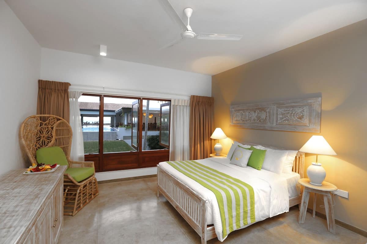 Ein Schlafzimmer mit einem Bett, zwei Nachttischlampen, einem Sessel, einem Tisch mit Obst und Blick auf einen Pool und ein Haus durch eine Schiebetür.