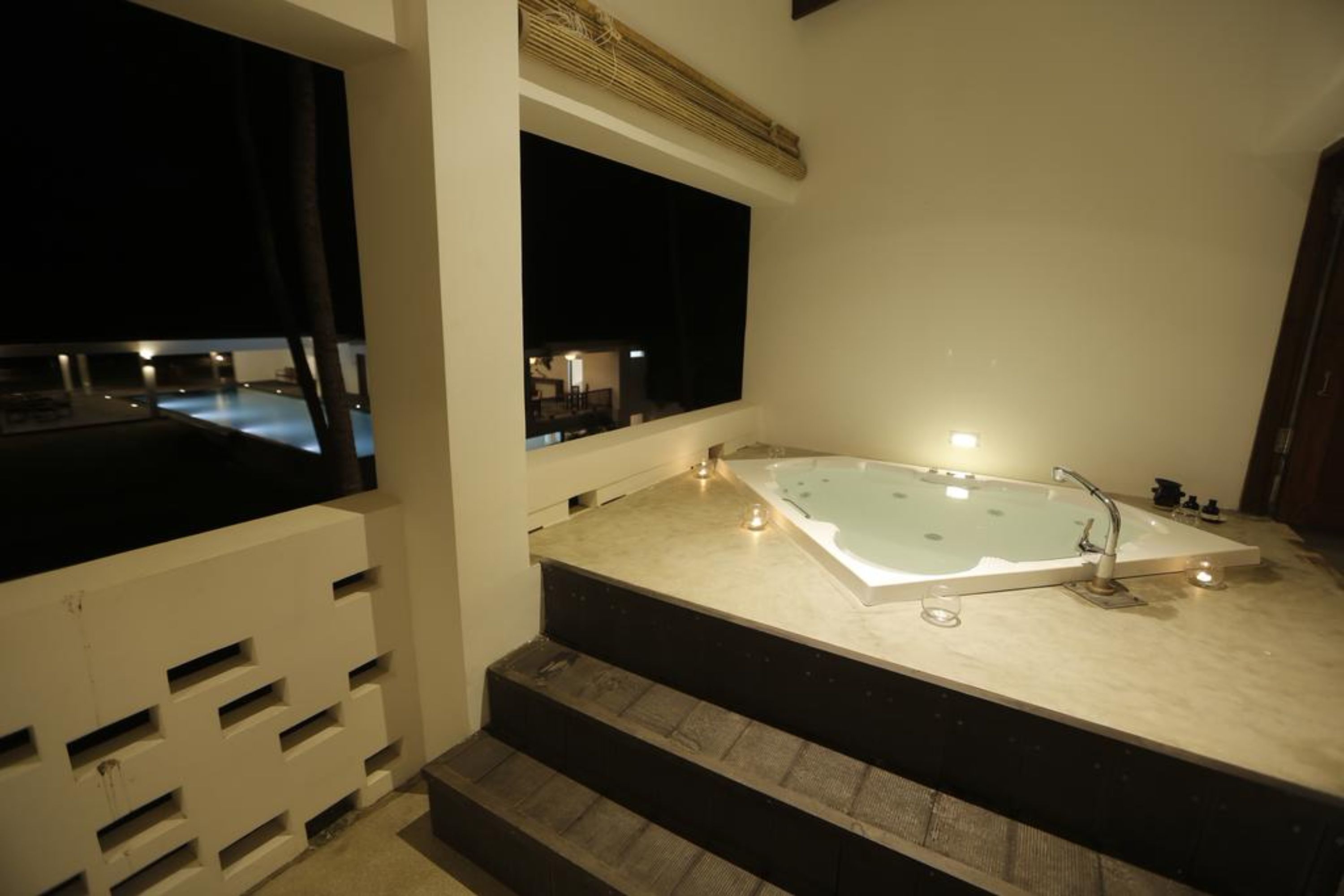 A modern bathroom with a large bathtub, a window overlooking a pool, and a staircase leading to a balcony.