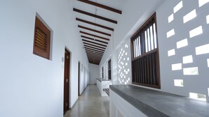 A long, narrow hallway with white walls and wooden beams on the ceiling. There are windows with wooden frames on one side and a patterned wall on the other. - The Beach Boutique Hotel (Uswetakeiyawa)