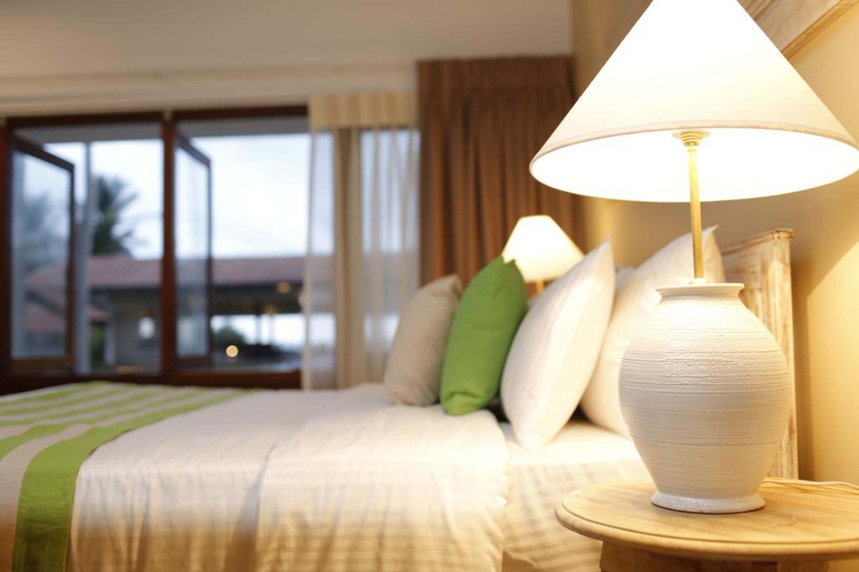 Una habitación de hotel con una cama, almohadas, una lámpara y un florero sobre una mesita de noche.