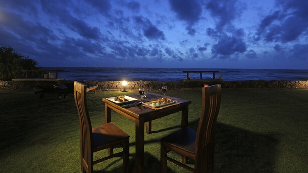 An outdoor dining table with chairs, a candle, and a bottle of wine, set against a backdrop of a beach and ocean under a cloudy sky. - The Beach Boutique Hotel (Uswetakeiyawa)