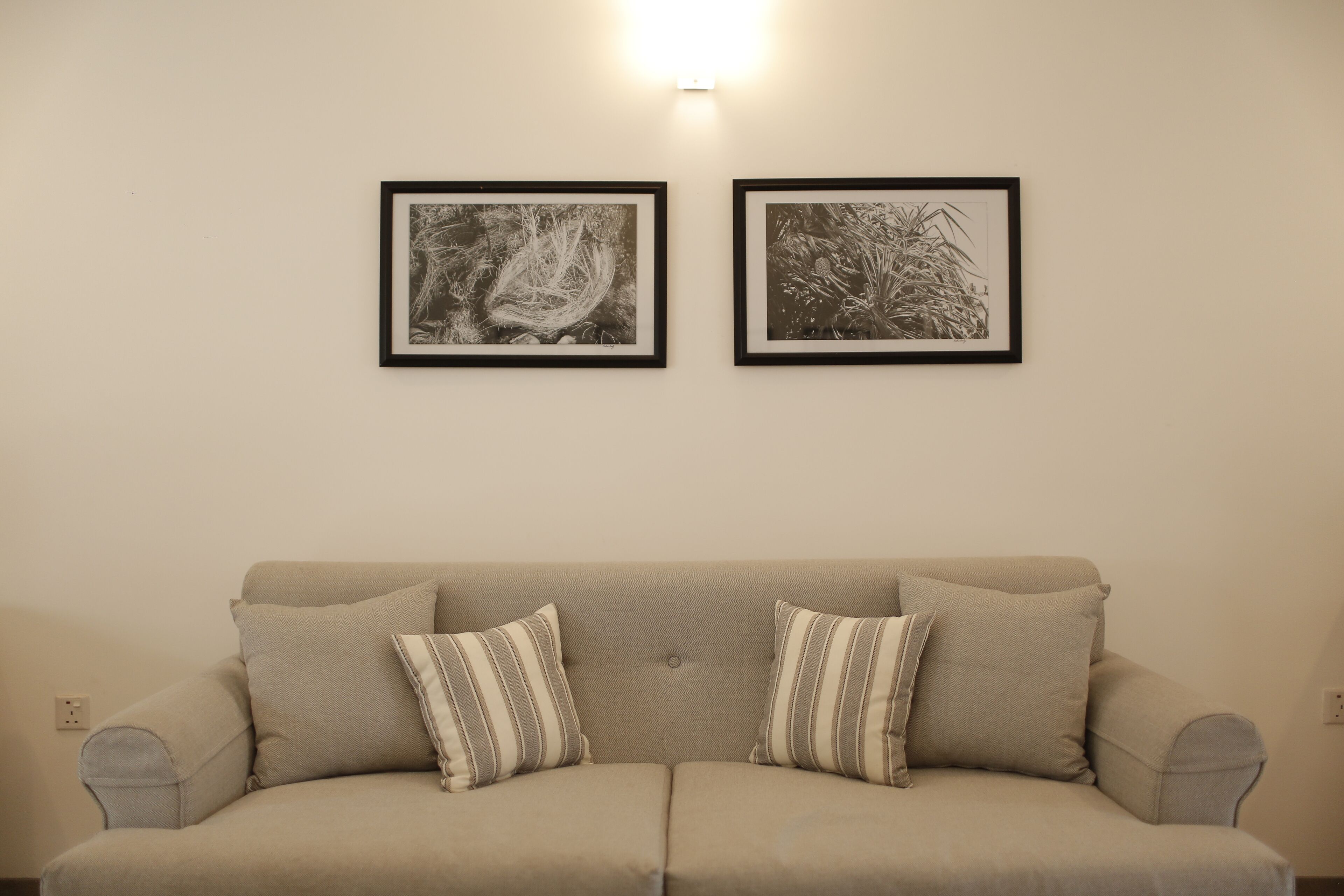 Un canapé beige agrémenté de coussins à rayures, deux photographies en noir et blanc encadrées accrochées au mur et un luminaire fixé au mur.