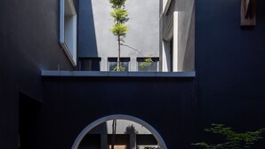 Una ventana circular con un pequeño árbol y dos luces esféricas en su interior, sobre un oscuro muro de edificio.