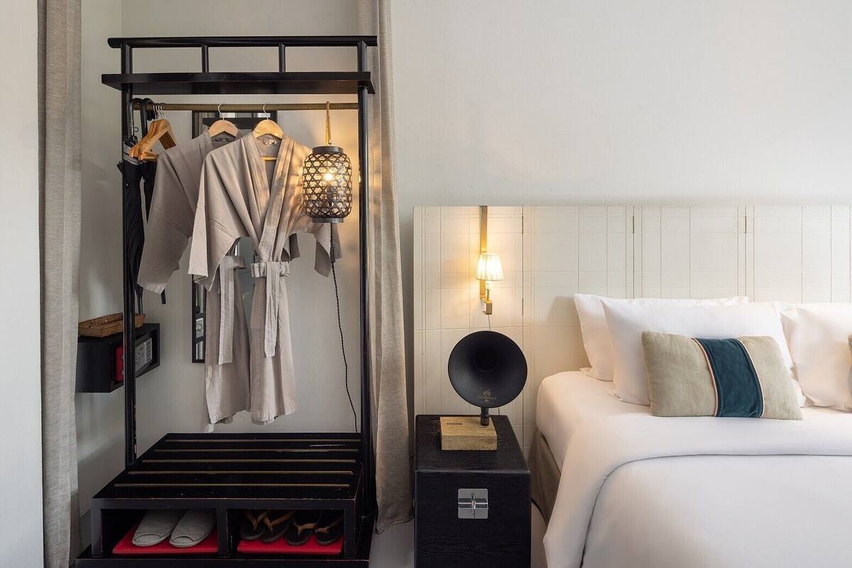 Una habitación de hotel con una cama, una mesita de noche con lámpara, un zapatero y un perchero con batas.