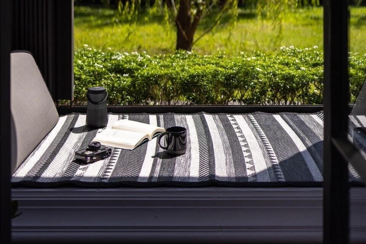 Asiento junto a la ventana con vistas a un jardín arbolado, un cojín a rayas blancas y negras, una cámara y un libro.