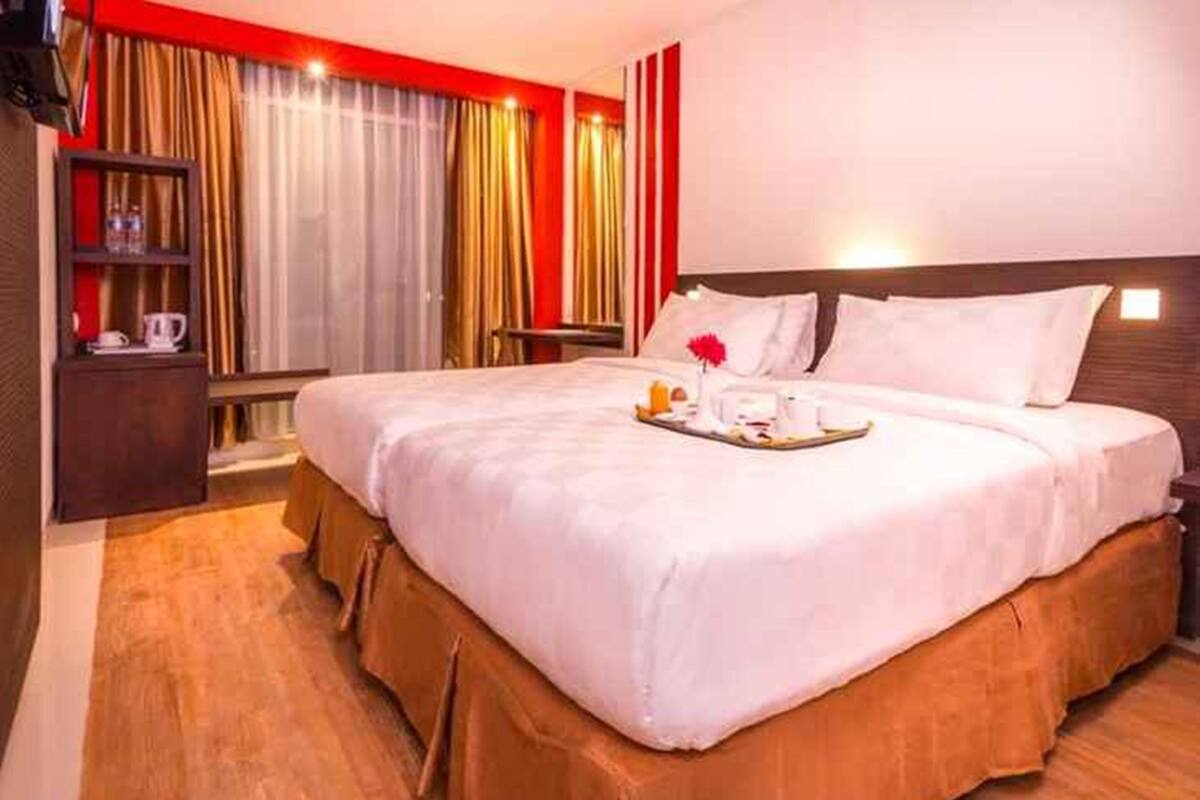 A hotel room with a large bed, a bedside table, a nightstand, and a shelf with a lamp and a book.