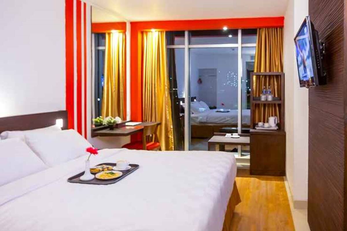 A hotel room with a large bed, a desk, a chair, a TV, and a sliding glass door leading to a balcony.