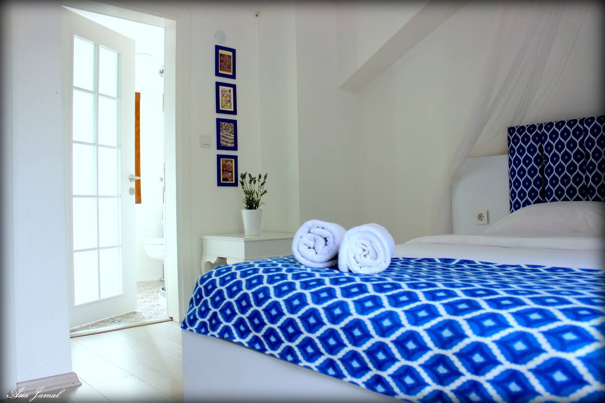 A hotel room with a bed, a small bedside table with a plant, and a door leading to a bathroom.
