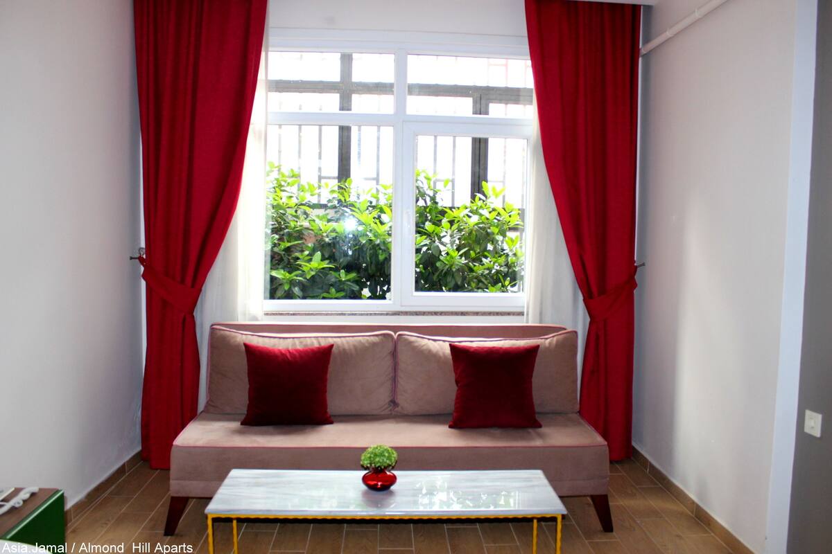 A room with a beige sofa, red curtains, a small table with a plant, and a window with a view of greenery.