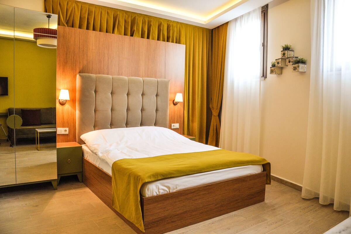 A hotel room with a wooden headboard, a bed with a white and yellow bedspread, a nightstand with a lamp, a mirror, and a wall-mounted shelf with plants.